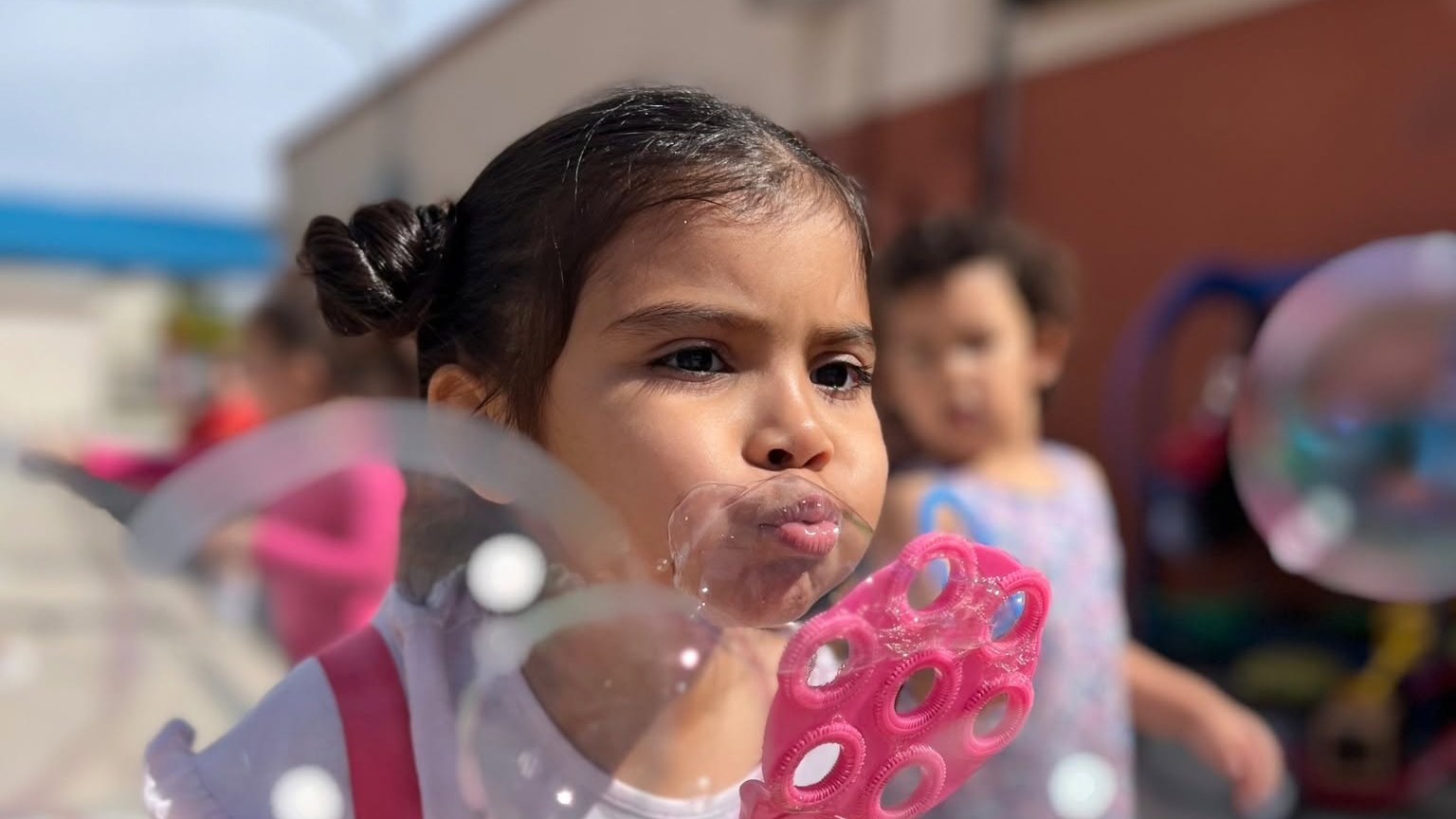 A girl blowing bubbles outside, with a classmate behind her